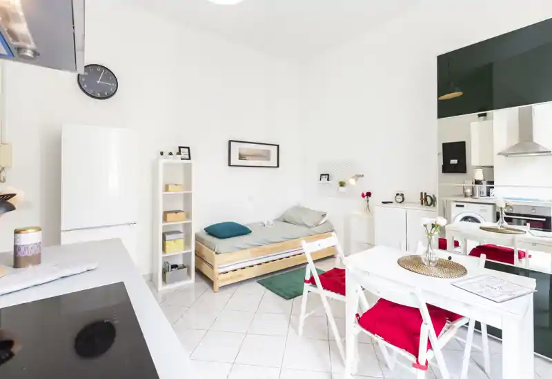 Monolocale luminoso con letto singolo a parete, angolo cottura compatto e piccolo tavolo da pranzo. Arredamento bianco e pulito con tocchi di colore su cuscini e tappeto.