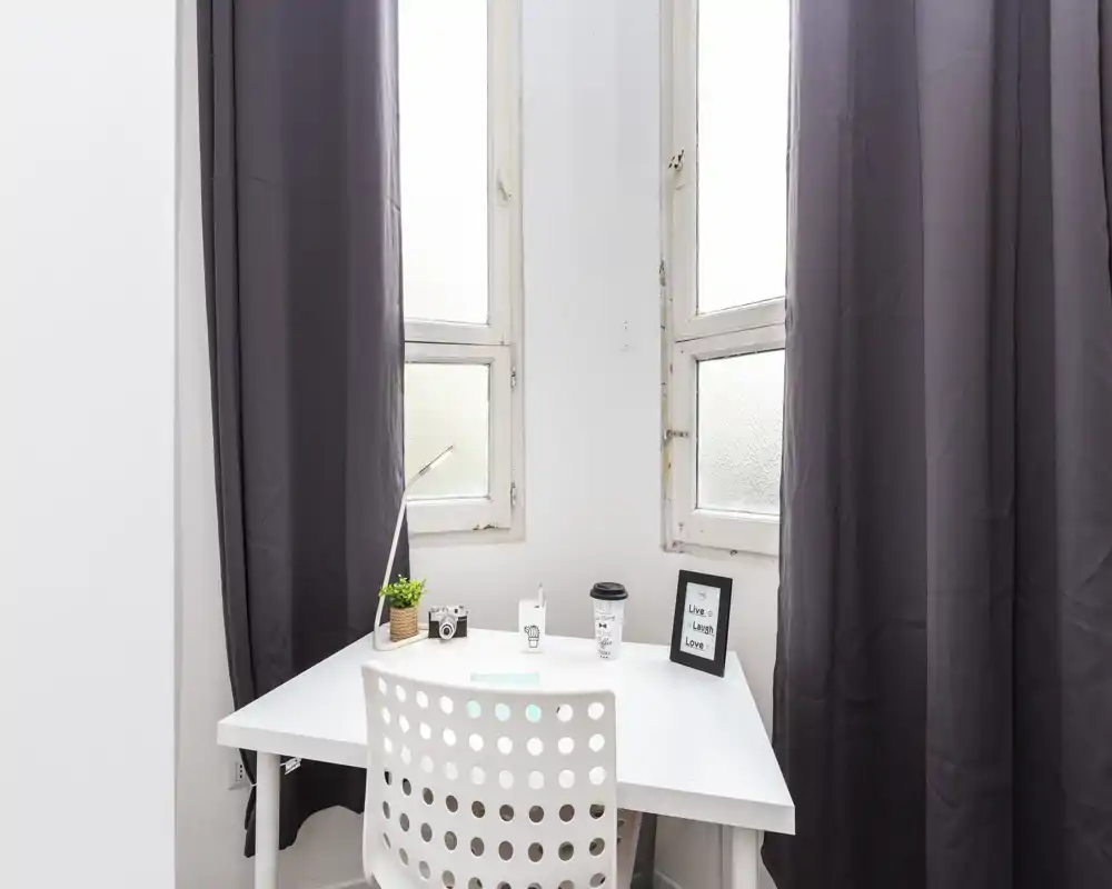 Bright, minimal workspace by two tall windows with a white desk, perforated chair and simple decorative items—ideal for a compact home office or study corner.