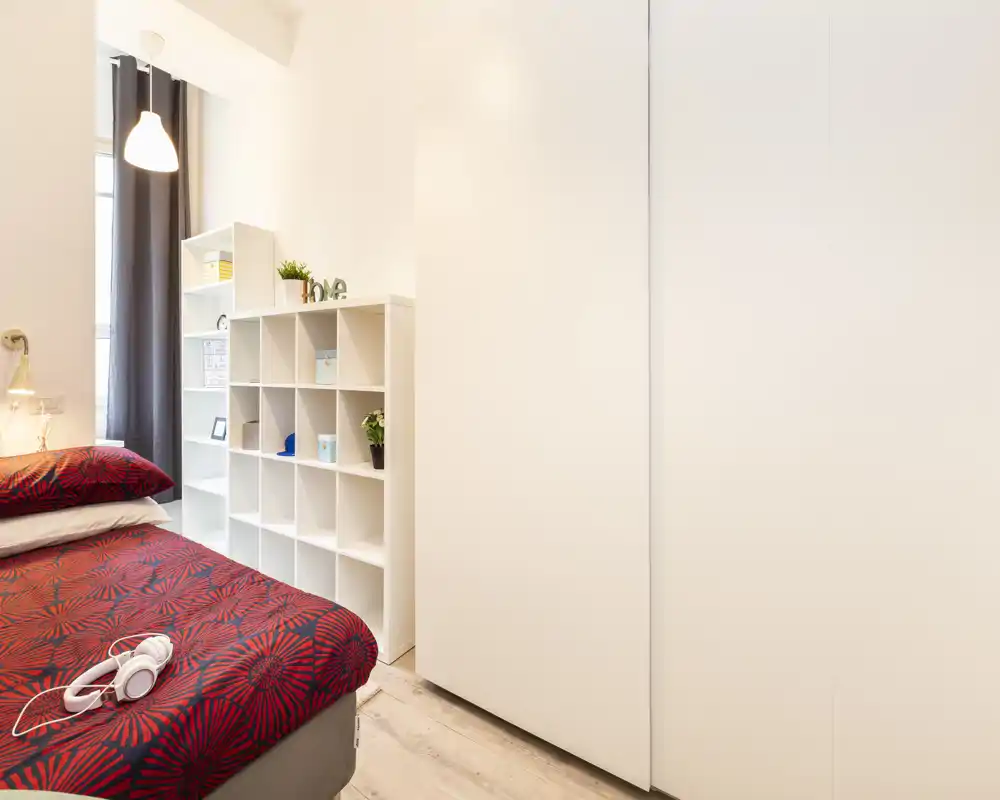 Bright, modern bedroom with a red patterned bedspread, built-in wardrobe and open shelving — neat, well-lit and welcoming.