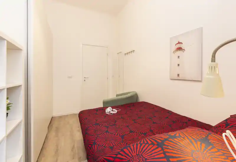 Bright, neatly styled bedroom with a bold red patterned bedspread, bedside lamp and minimal decor — ideal for a cozy single or double stay.