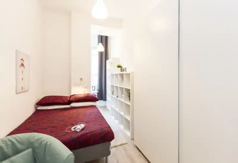 Bright, modern single bedroom with a red patterned bedspread, bedside lamp and shelving unit; clean, minimal styling ideal for short-term rentals.