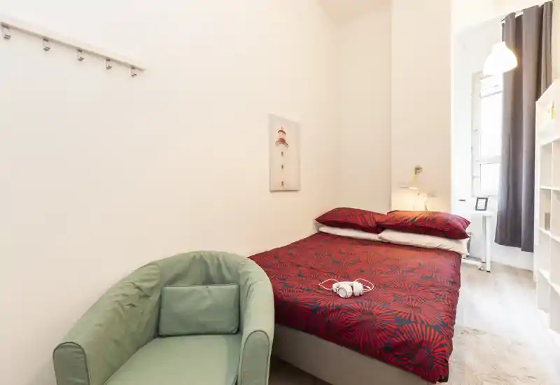 Bright, compact bedroom with a double bed dressed in patterned red linens, a bedside lamp and a comfortable green armchair by the wall. Natural light enters from a nearby window; the space is neatly styled and inviting.