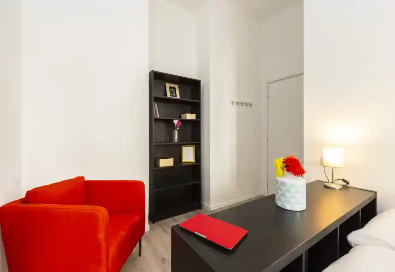Zona giorno luminosa e moderna con una poltrona rossa vivace, libreria e piano d'appoggio con fiori decorativi e lampada — un angolo accogliente e ben illuminato, ideale per rilassarsi o lavorare.