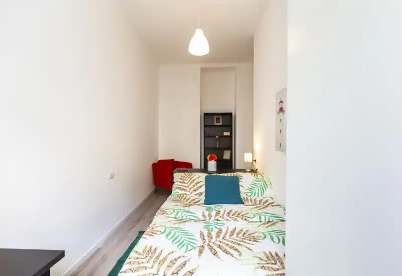 Dormitorio compacto y luminoso con ropa de cama estampada, cojín turquesa y lámpara de noche; un sillón rojo y una estantería al fondo crean un ambiente acogedor y con estilo.