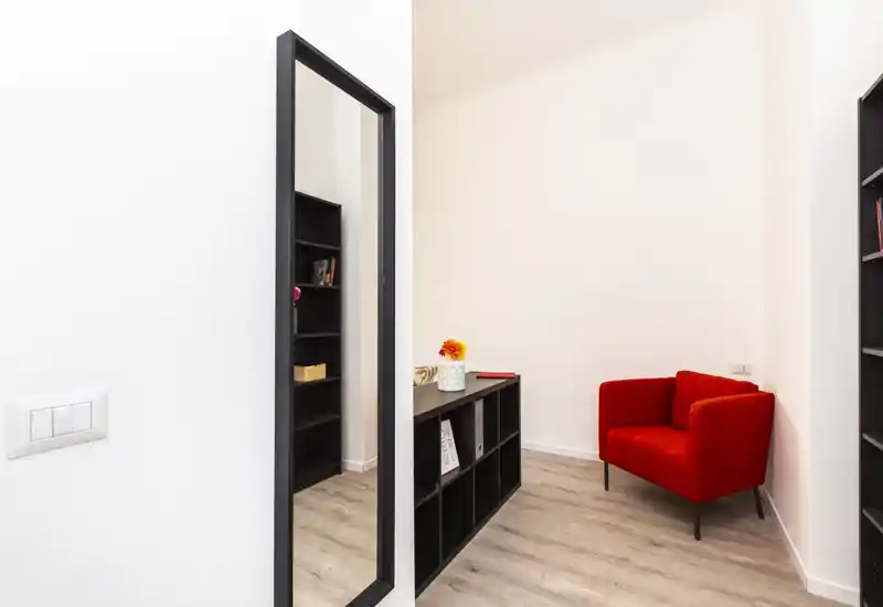 Angolo living luminoso e compatto con una poltrona rossa vivace, scaffalature sottili e uno specchio alto — stile minimal e moderno.