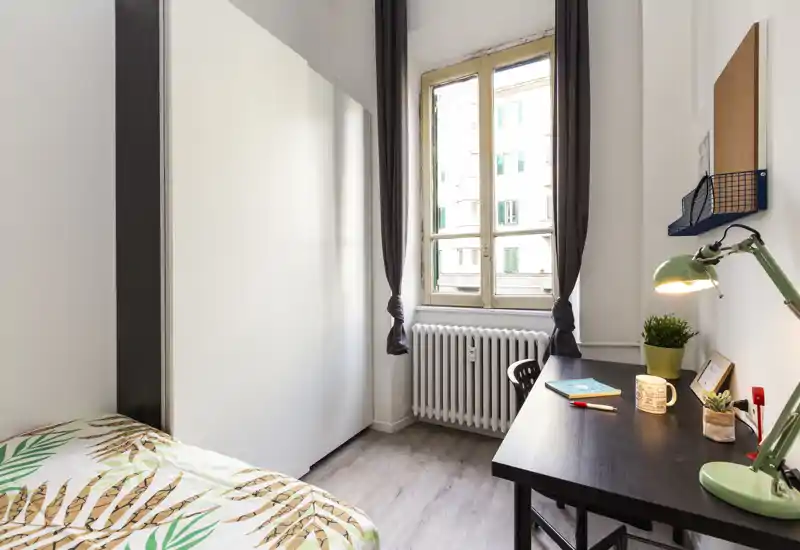 Habitación compacta y bien iluminada con cama individual, armario y escritorio ordenado junto a la ventana—ideal para estudiante o una sola persona.