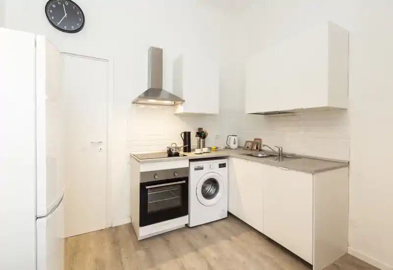 Cucina moderna compatta con mobili bianchi, forno integrato, cappa, lavello e lavatrice — luminosa, pulita e organizzata in modo efficiente per spazi ridotti.
