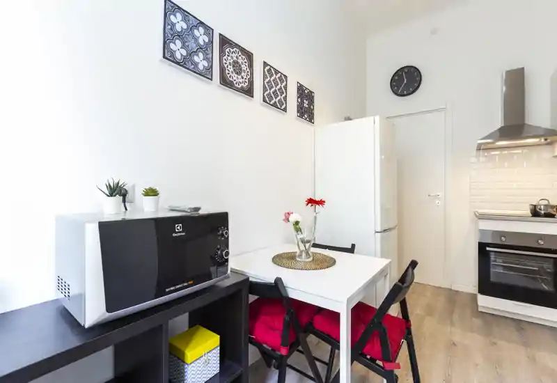Cucina compatta e luminosa con forno moderno, frigorifero e un piccolo tavolo da pranzo. Ben arredata con piastrelle decorative e un vaso di fiori per un tocco accogliente.