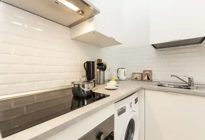 Cucina moderna compatta con piano cottura in vetroceramica, lavello in acciaio e piccoli elettrodomestici sul piano. Il paraschizzi a mattonelle bianche e l'arredo curato rendono l'ambiente luminoso e accogliente.