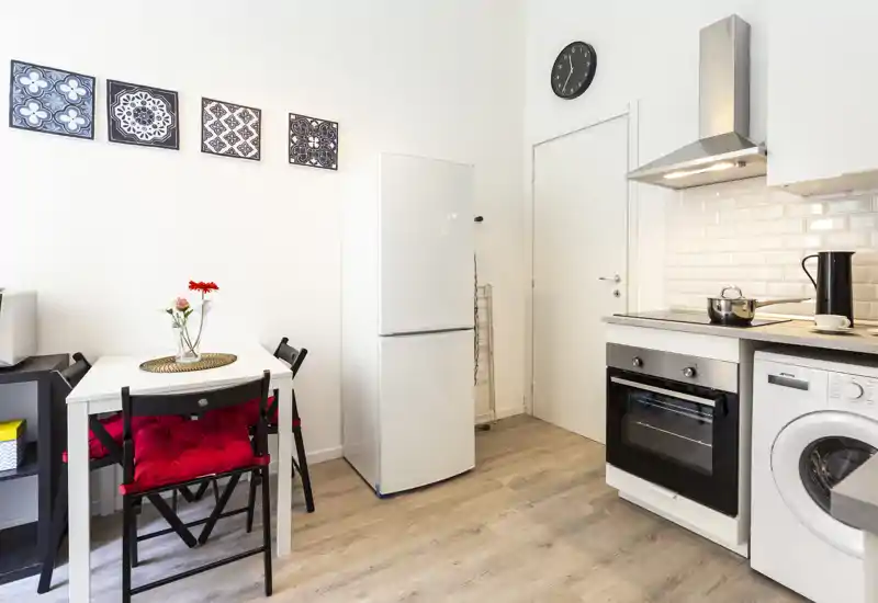 Cucina moderna compatta con frigorifero, forno e piccolo tavolo da pranzo con sedie con cuscini rossi; ambiente luminoso e arredato con cura, ideale per affitti brevi.