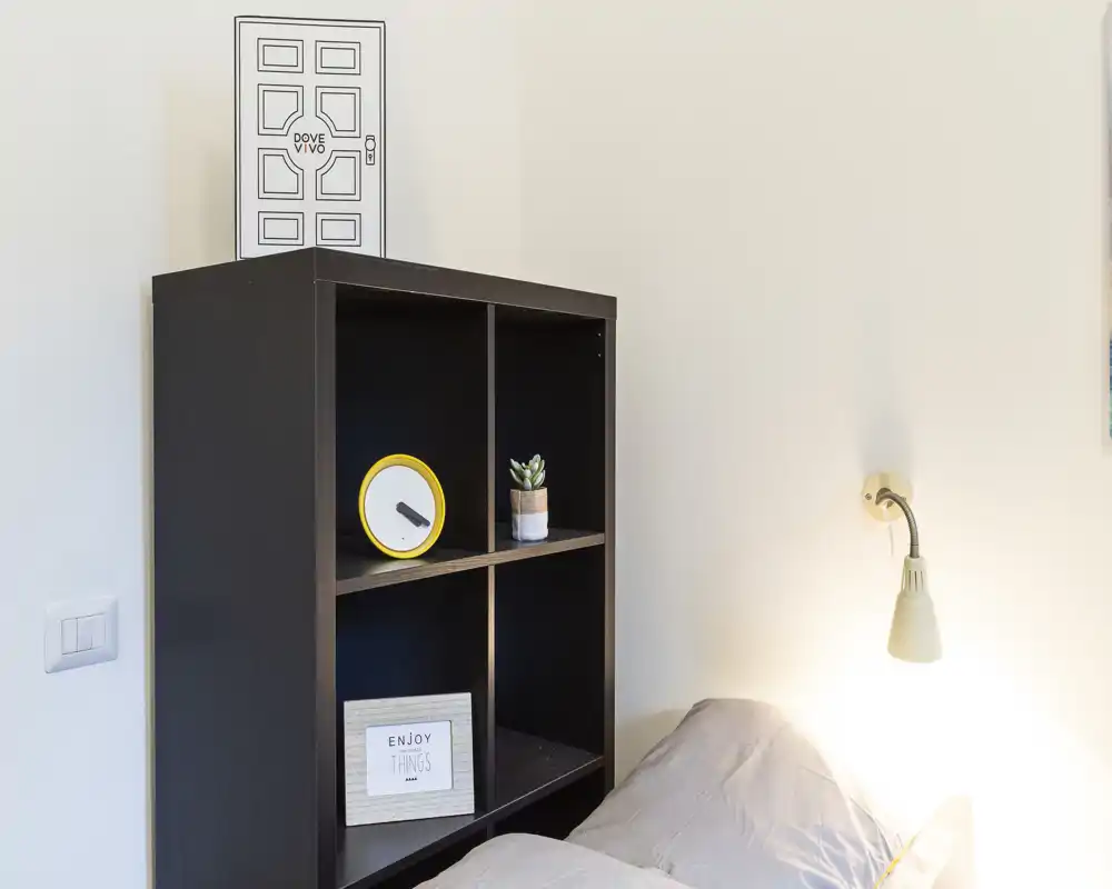 Angolo di camera con scaffale accanto al letto, parte del materasso con cuscini e una lampada a muro — ordinato, illuminazione soffusa e arredo essenziale.