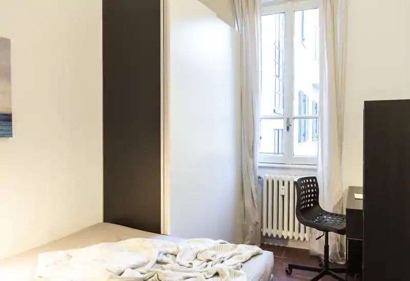 Camera da letto minimalista con letto matrimoniale visibile, luce naturale da una finestra alta e un angolo studio compatto con sedia e cassettiera.