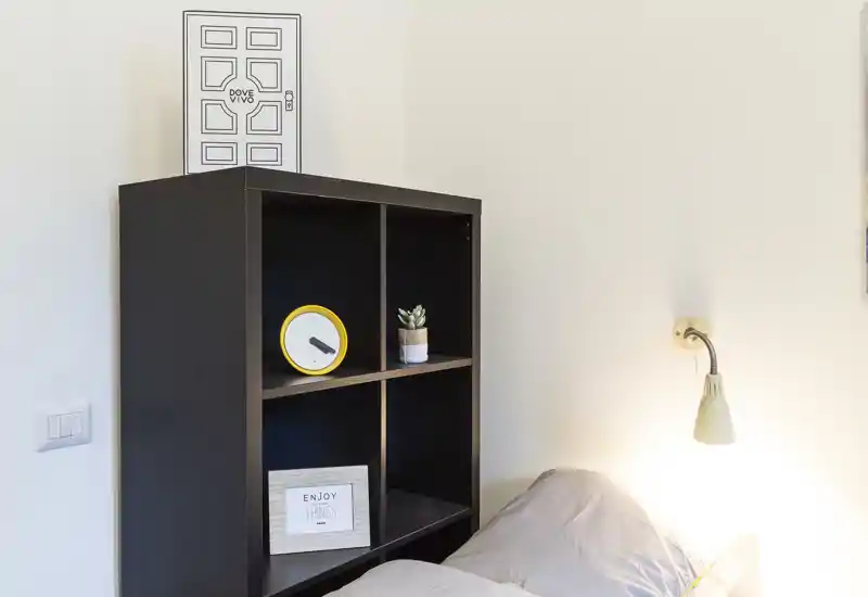 Angolo di camera con scaffale accanto al letto, parte del materasso con cuscini e una lampada a muro — ordinato, illuminazione soffusa e arredo essenziale.