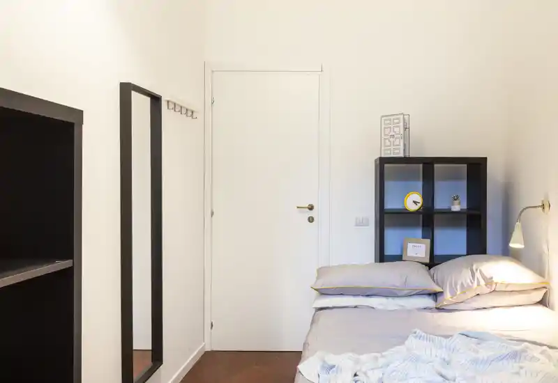 Camera da letto accogliente e ben illuminata con letto ordinato, mensola laterale e lampada a parete — perfetta per una persona o brevi soggiorni.