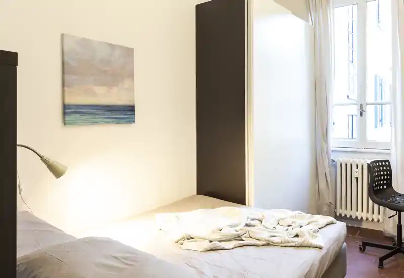 Camera da letto illuminata dalla luce naturale con un letto matrimoniale, lenzuola morbide e una lampada da comodino. Un armadio e una finestra con tende completano l'ambiente semplice e confortevole.