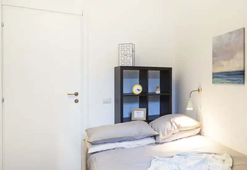 Angolo di camera da letto luminoso e moderno con letto matrimoniale ben rifatto, lampada da comodino e scaffale a cubi per riporre oggetti — toni neutri e quadro con tema marino per un’atmosfera rilassante.