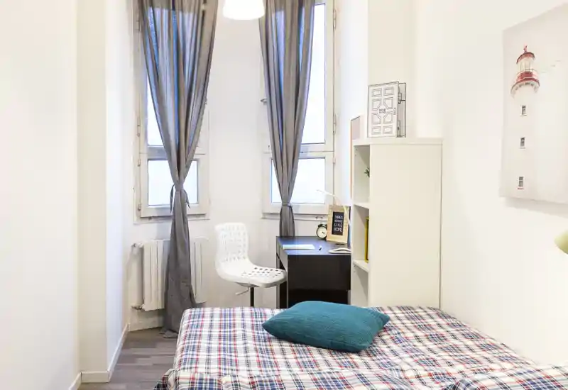 Chambre lumineuse et compacte avec lit soigné, coussin turquoise et un petit bureau près de grandes fenêtres laissant entrer la lumière naturelle.