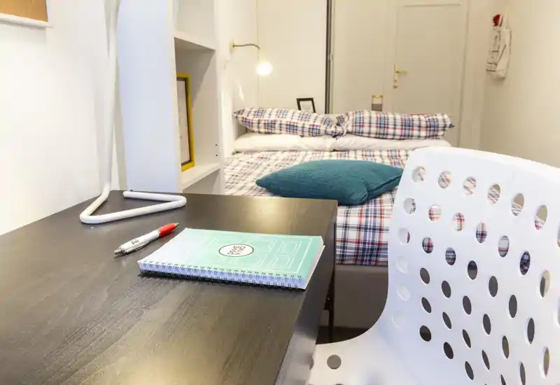 Chambre simple chaleureuse et lumineuse avec un bureau au premier plan, linge à carreaux et coussin décoratif — idéale pour étudiant ou location courte durée.