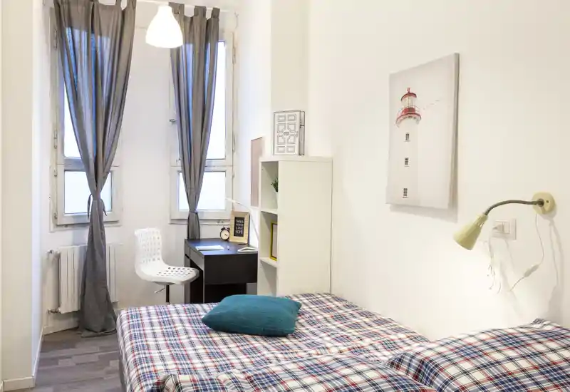 Chambre compacte et lumineuse avec un lit habillé de housses à carreaux, un coussin turquoise et un petit coin bureau près de la fenêtre. Murs neutres, affiche de phare et éclairage doux pour une atmosphère propre et accueillante.