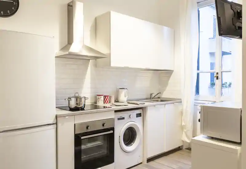 Cucina moderna compatta con mobili bianchi, forno integrato, lavatrice e una finestra luminosa che fornisce luce naturale — ordinata e pronta all'uso.