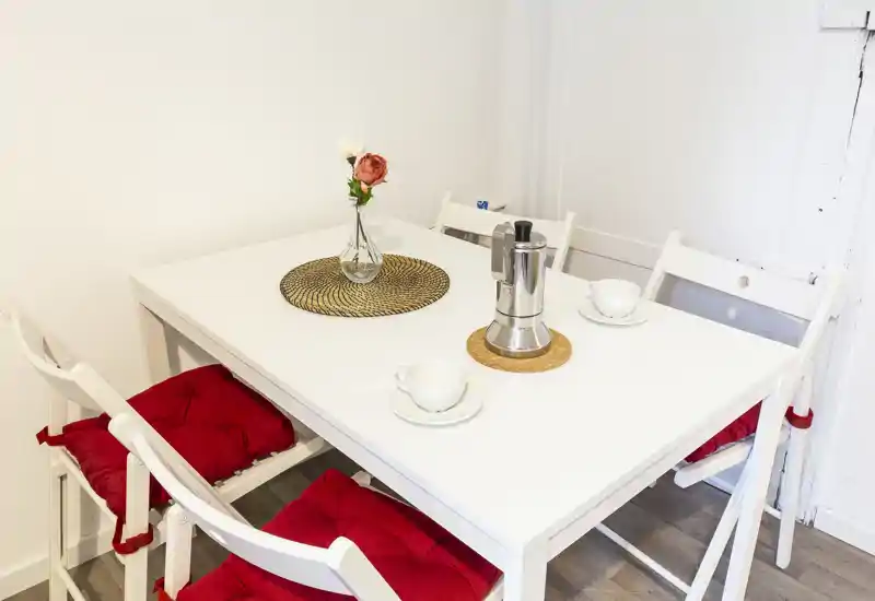 Zona pranzo luminosa e minimal con tavolo bianco, quattro sedie pieghevoli con cuscini rossi, un piccolo vaso con fiori e tazze da caffè — perfetta per pasti informali.