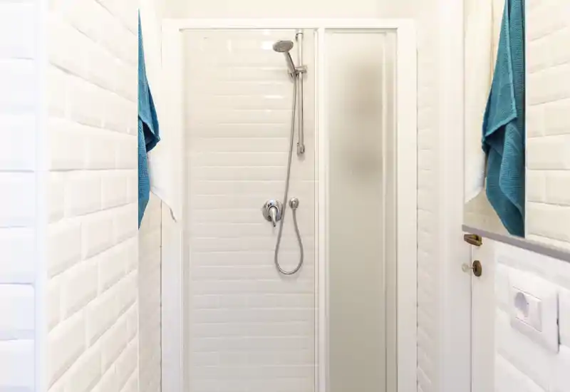 Doccia compatta e luminosa con piastrelle bianche effetto subway, soffione cromato con doccetta e porta doccia in vetro satinato; asciugamani turchesi per un tocco fresco.