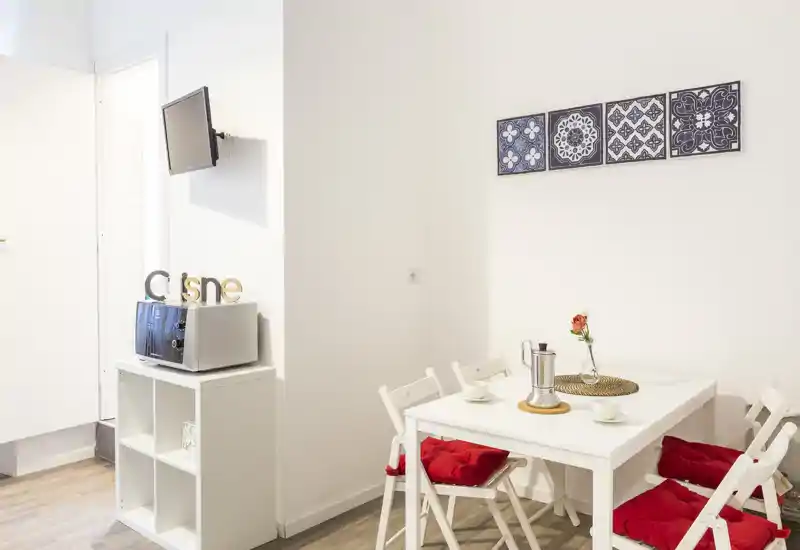 Angolo pranzo luminoso e compatto con tavolo bianco, quattro sedie con cuscini rossi; decorazioni semplici includono un piccolo fiore e piastrelle a parete, ideale per pasti accoglienti.