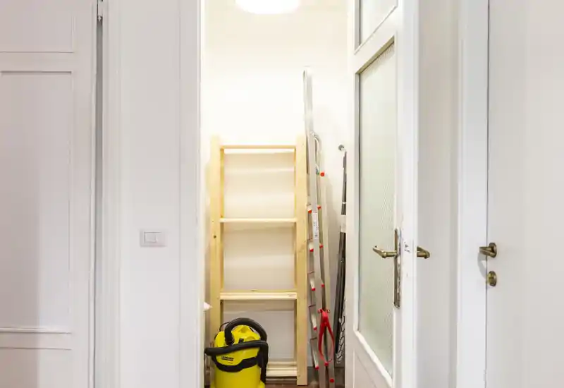 Piccolo vano d'ingresso/utility con una scaffalatura, una scala e un aspirapolvere industriale giallo. Pareti bianche neutre e una porta con pannello in vetro delimitano lo spazio di deposito.
