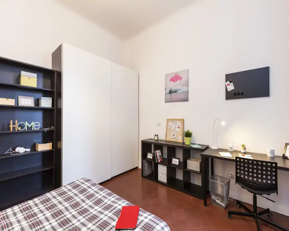 Chambre lumineuse et bien mise en scène avec lit à motifs au premier plan, grand placard, étagères et bureau rangé — idéale pour un étudiant ou une personne seule.