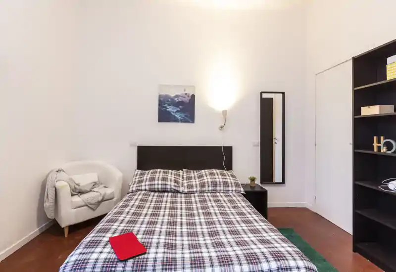 Chambre lumineuse et soignée avec lit double recouvert d'une couette à carreaux, un fauteuil confortable, table de chevet avec lampe et grand miroir — idéale pour un séjour agréable.