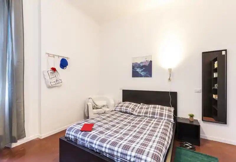 Chambre lumineuse et confortable avec un lit double en parure à carreaux, une table de chevet avec lampe, un petit fauteuil et une grande glace — parfaite pour un séjour cosy.