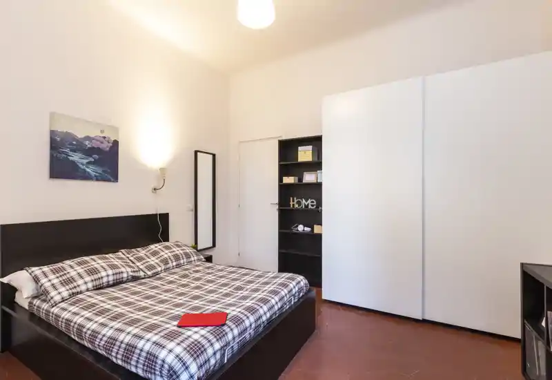 Chambre claire et bien rangée avec grand lit double, placard et étagères. L'éclairage doux et la literie à carreaux offrent un espace de sommeil accueillant et lumineux.