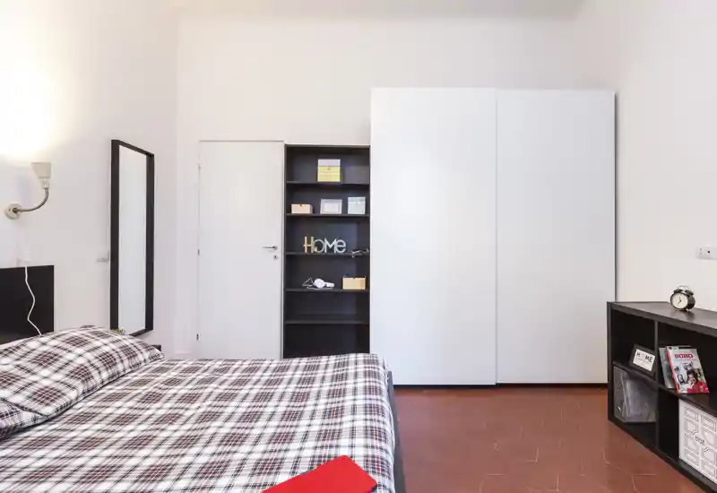 Chambre lumineuse et bien rangée avec un grand lit au premier plan, une literie à carreaux, une armoire et des étagères de rangement. Lignes épurées et éclairage doux, idéal pour une annonce locative.