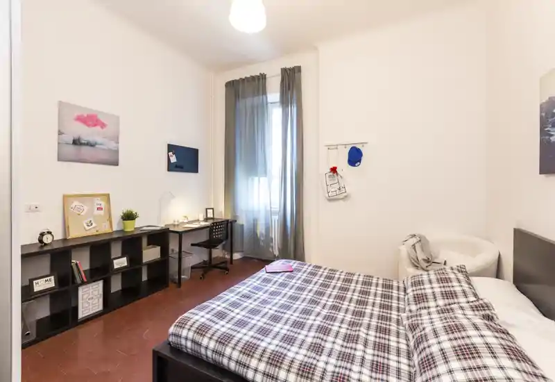 Chambre claire et chaleureuse avec lit double en couvre-lit à carreaux, un bureau près de la fenêtre, étagères et un fauteuil cosy — idéale pour étudiants ou professionnels.