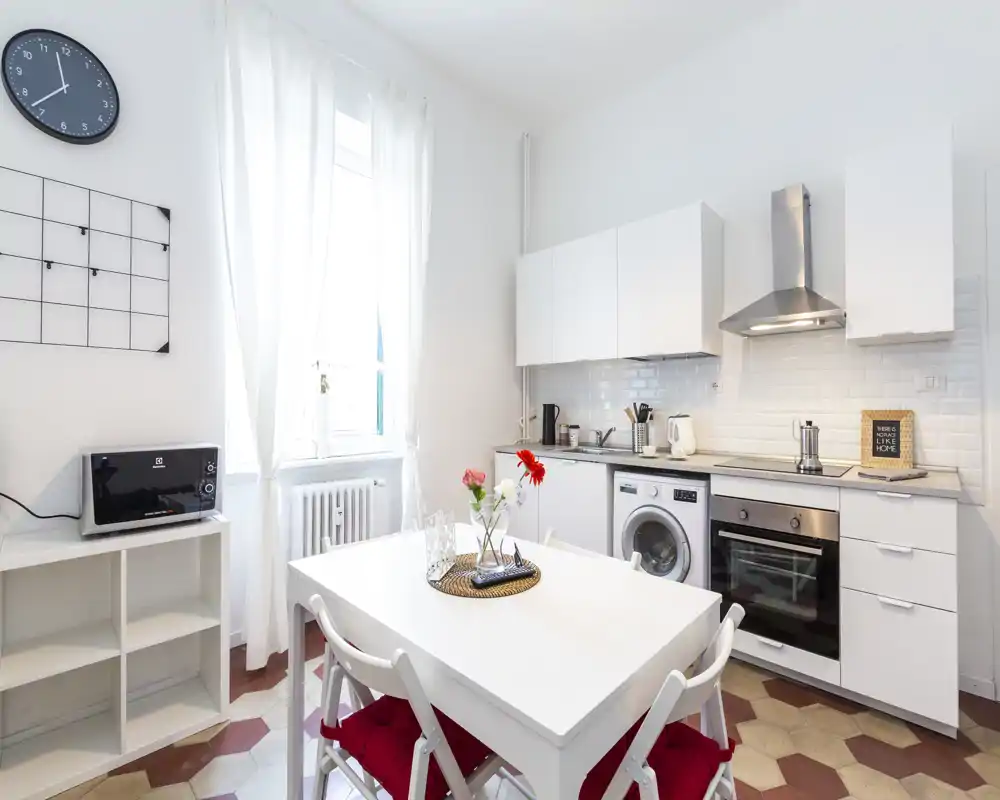 Cuisine moderne et lumineuse avec électroménagers intégrés et table centrale. Placards blancs, coin repas chaleureux avec chaises à coussin rouge et décoration florale.