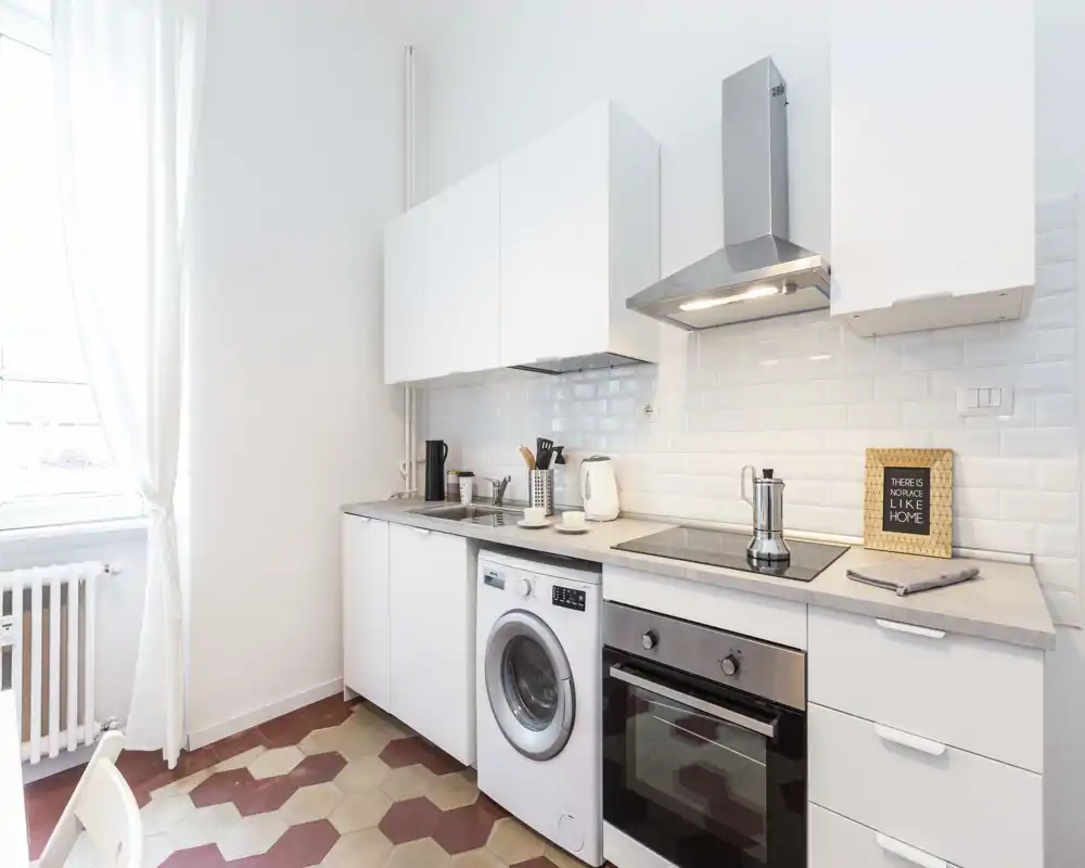 Coin cuisine moderne et lumineux avec meubles blancs, four encastré, plaque à induction et machine à laver sous le plan de travail. La lumière naturelle et la crédence en carreaux métro donnent une atmosphère aérée et pratique.