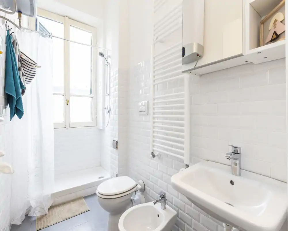 Salle de bains lumineuse et propre avec douche, lavabo, bidet et WC ; carreaux blancs et lumière naturelle provenant d'une fenêtre pour une atmosphère fraîche.