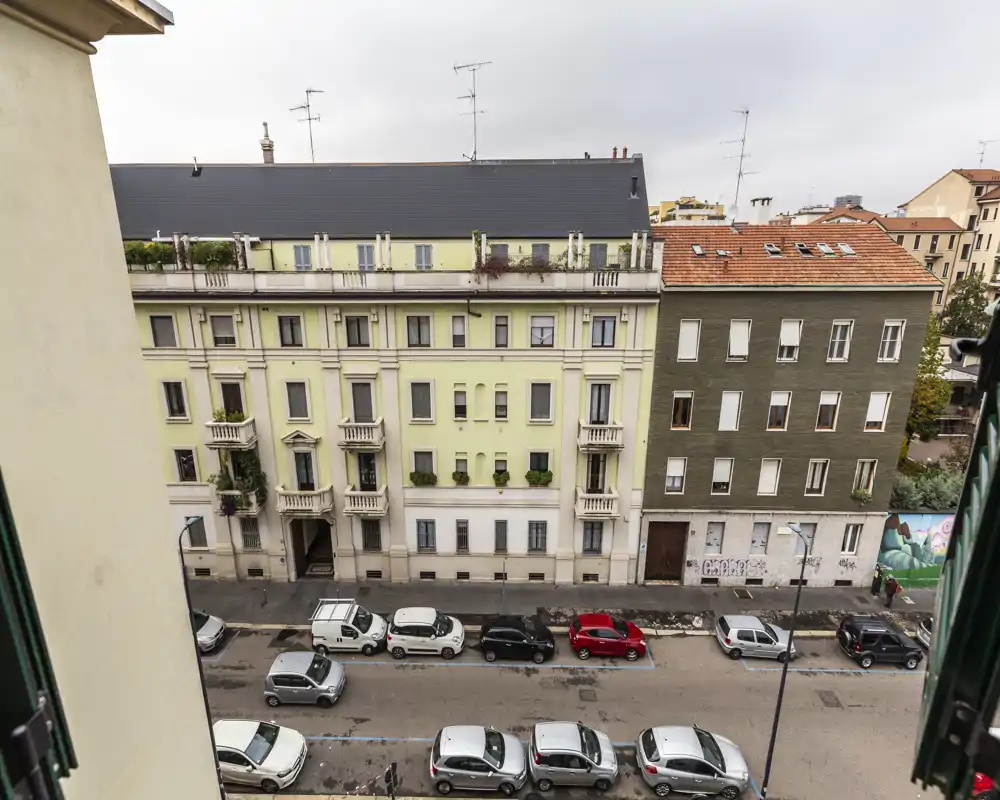 Vue sur la rue depuis une fenêtre ou un balcon à l'étage montrant des voitures garées et des immeubles résidentiels sous un ciel couvert.