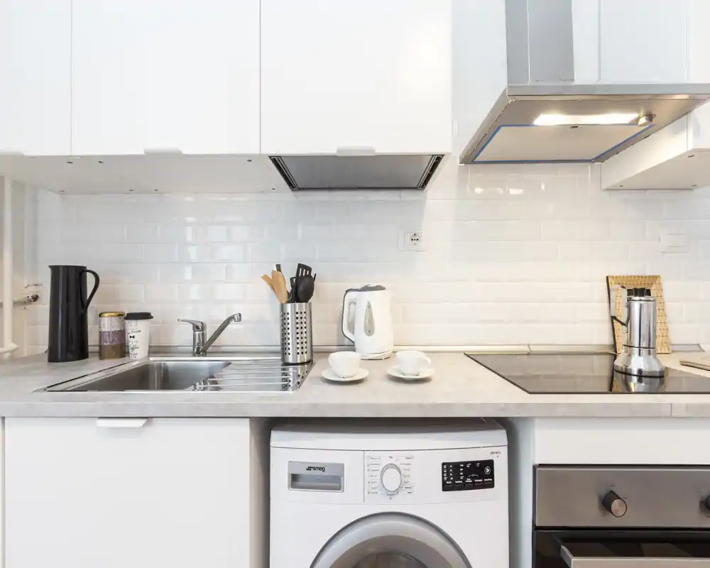 Cuisine compacte et moderne avec meubles blancs, évier inox, plaque à induction et petits appareils. Plan de travail propre et lumineux avec bouilloire, ustensiles et deux tasses.