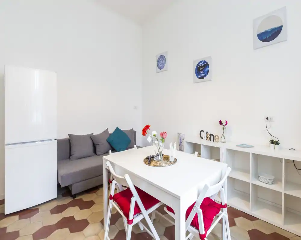 Espace de vie compact et lumineux avec table blanche, chaises pliantes à coussins rouges, petit canapé gris et réfrigérateur — espace moderne et soigné, parfait pour courts séjours.