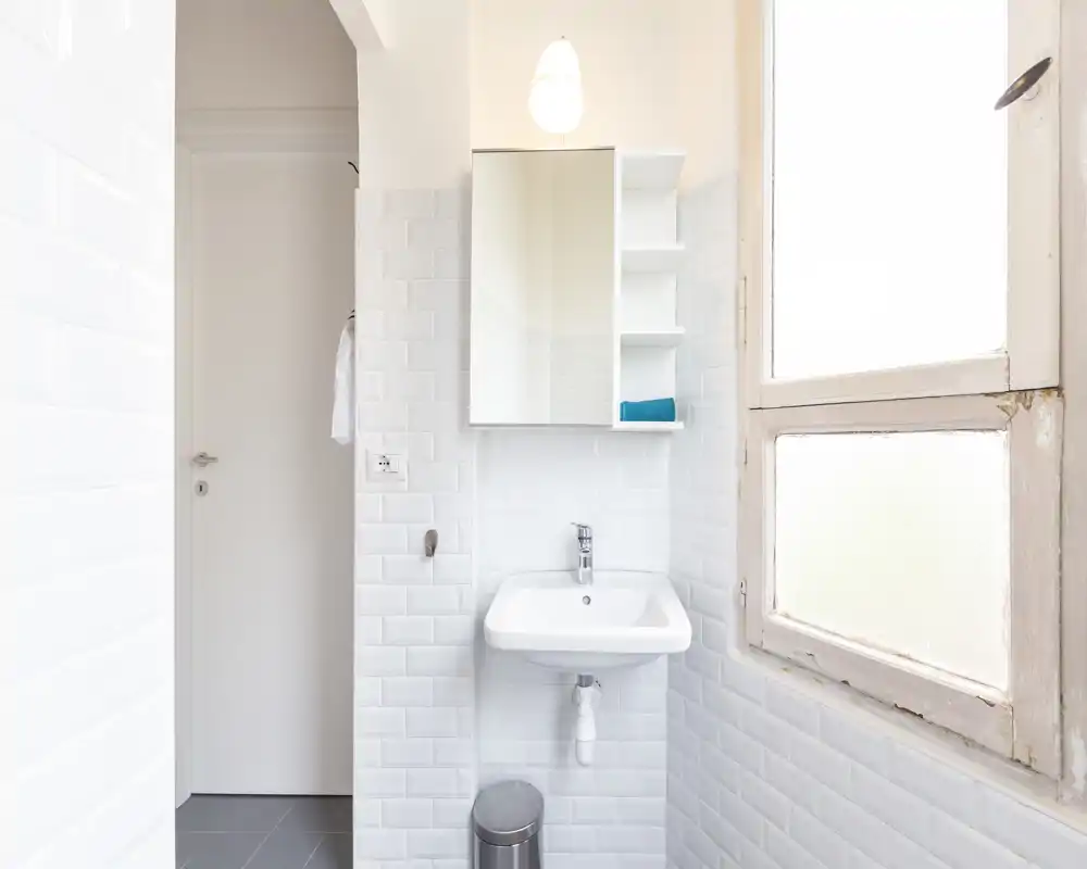 Salle de bain lumineuse et minimaliste avec lavabo mural, armoire-miroir et murs carrelés. Une grande fenêtre dépoli laisse entrer la lumière naturelle, créant une atmosphère propre et aérienne.