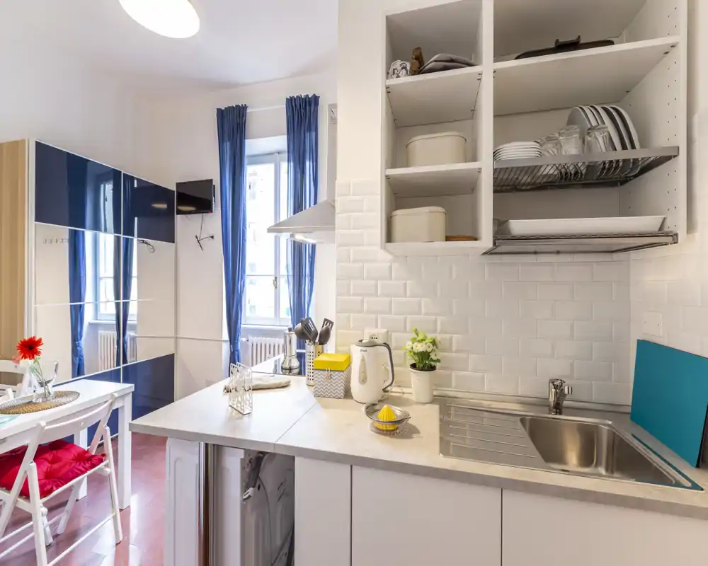 Kitchenette compacte et moderne avec évier en inox, étagères ouvertes et fenêtre lumineuse. Plan de travail propre, petits appareils et touches décoratives la rendent accueillante.