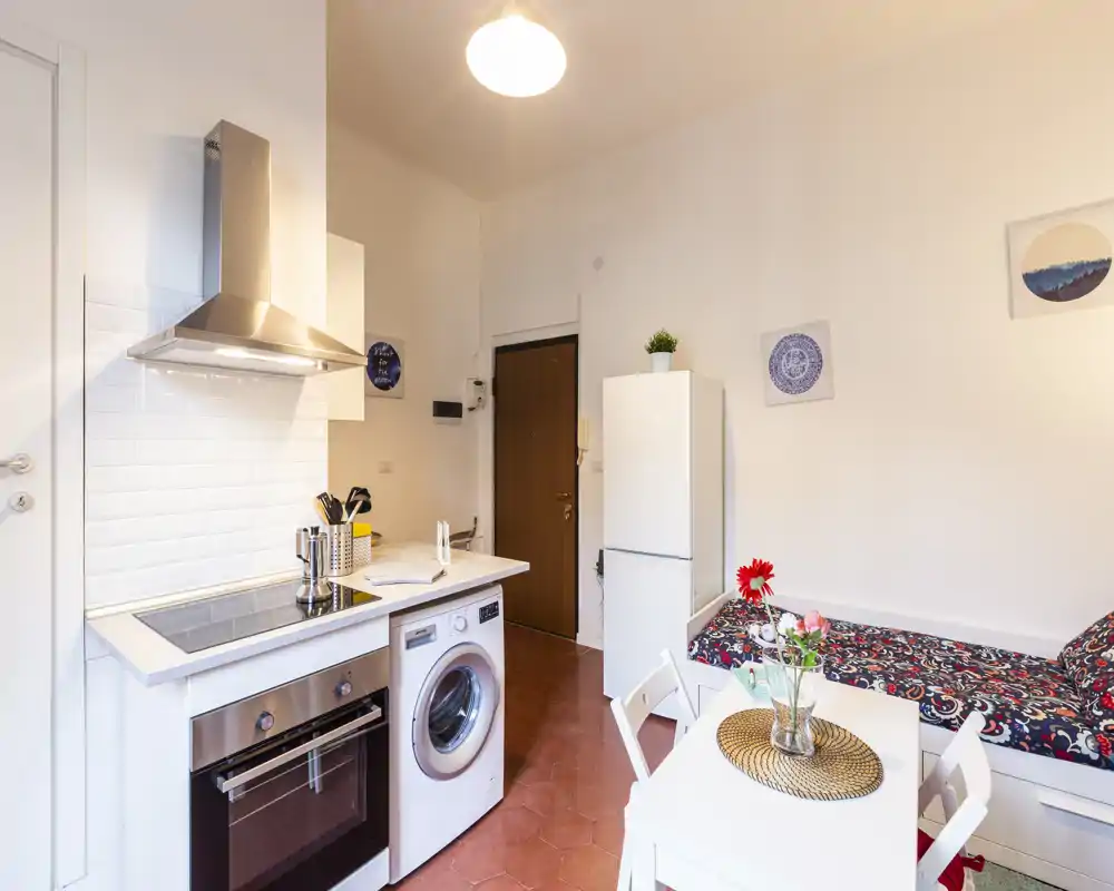 Cuisine compacte ouverte dans un studio, équipée d'une plaque à induction, d'un four encastré, d'une hotte et d'une machine à laver sous le plan de travail. Espace soigné avec une petite table, des fleurs décoratives et un lit-banquette sur le côté.