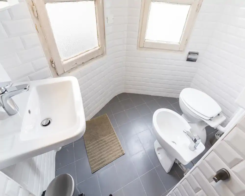 Salle de bain compacte et lumineuse avec murs carrelés blancs, lavabo mural, bidet et toilettes; sol gris et fenêtres dépoli pour intimité et lumière naturelle.