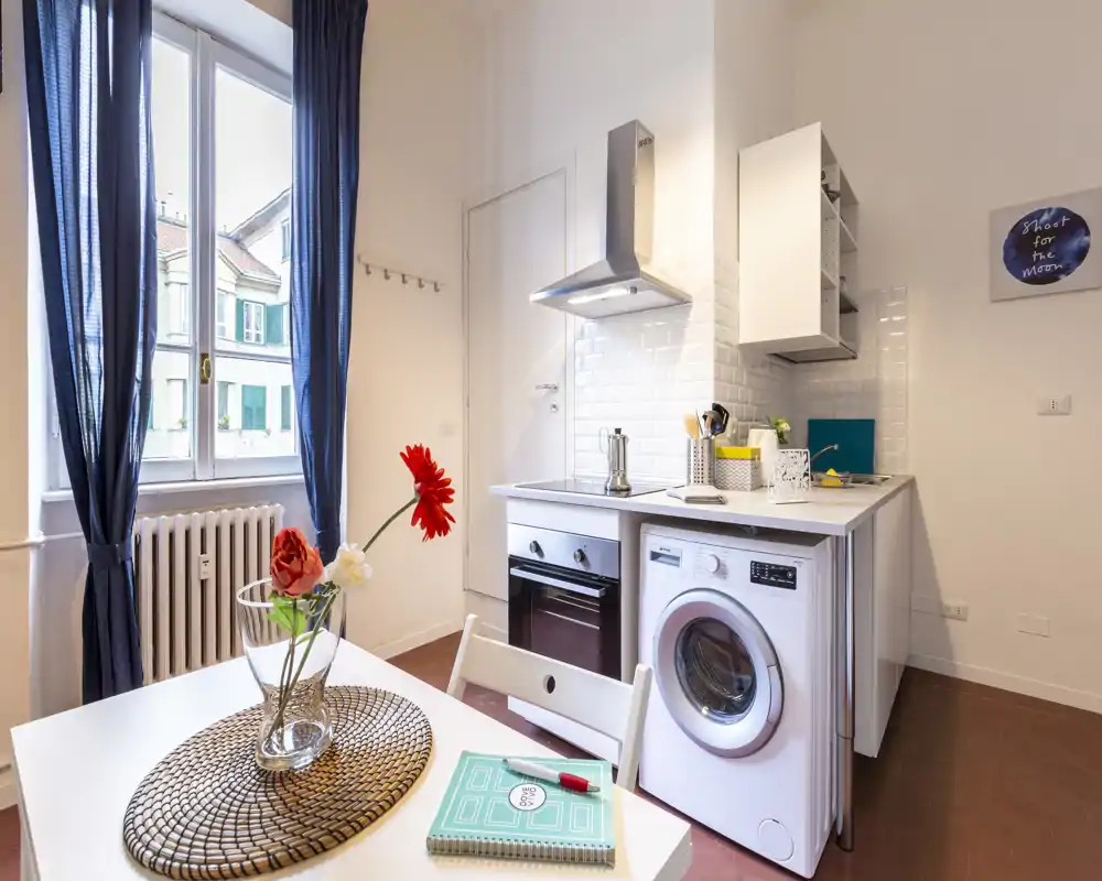 Kitchenette moderne et compacte avec four, plaque et lave-linge, fenêtre lumineuse et table soignée — idéale pour un studio en ville.