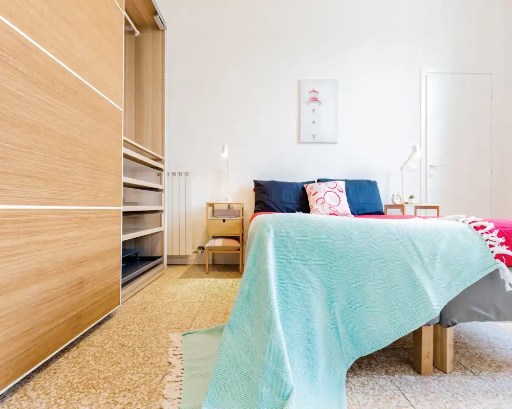 Chambre lumineuse et bien mise en scène avec un lit double recouvert de textiles clairs, des tables de chevet et des lampes, ainsi qu'une armoire encastrée. L'atmosphère est fraîche et accueillante, avec une lumière naturelle et des tons neutres.