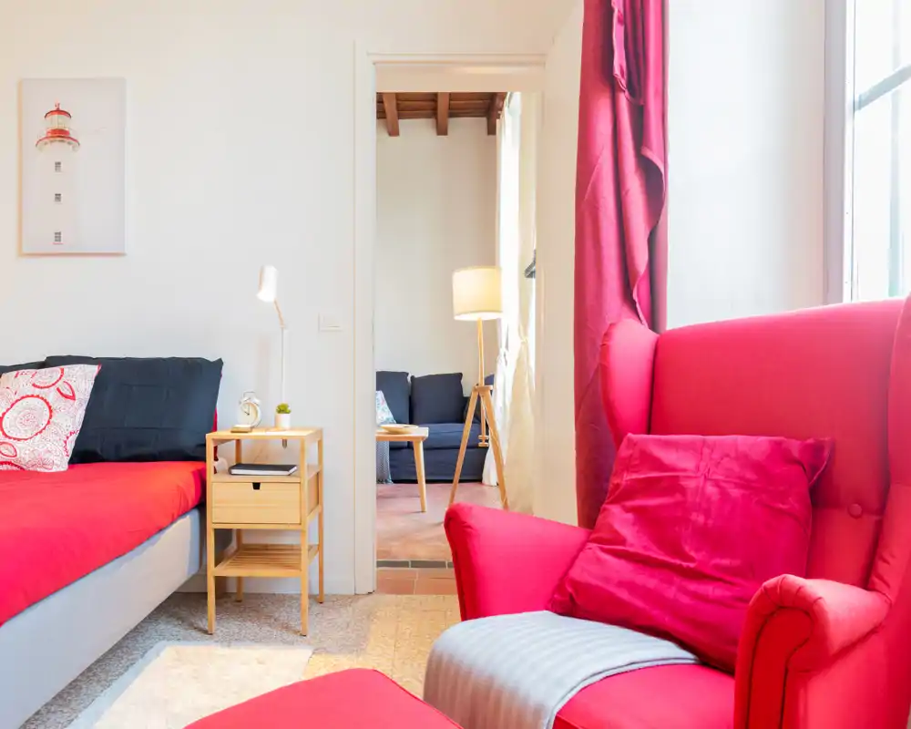 Coin chambre lumineux et chaleureux avec un fauteuil rouge et un lit bien fait orné de coussins; la lumière naturelle entre par la fenêtre et une porte laisse entrevoir un petit salon.