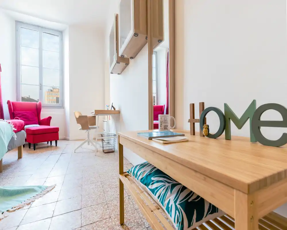 Chambre lumineuse et stylée avec un lit confortable, un fauteuil rouge près de la fenêtre et une console en bois avec des éléments décoratifs — idéale pour une annonce chaleureuse.