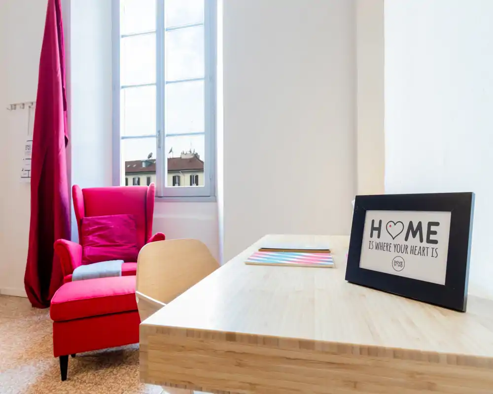 Coin salon lumineux et moderne avec un fauteuil rouge vif près d'une grande fenêtre et un bureau en bois minimaliste — espace chaleureux et bien éclairé, parfait pour se détendre ou travailler.