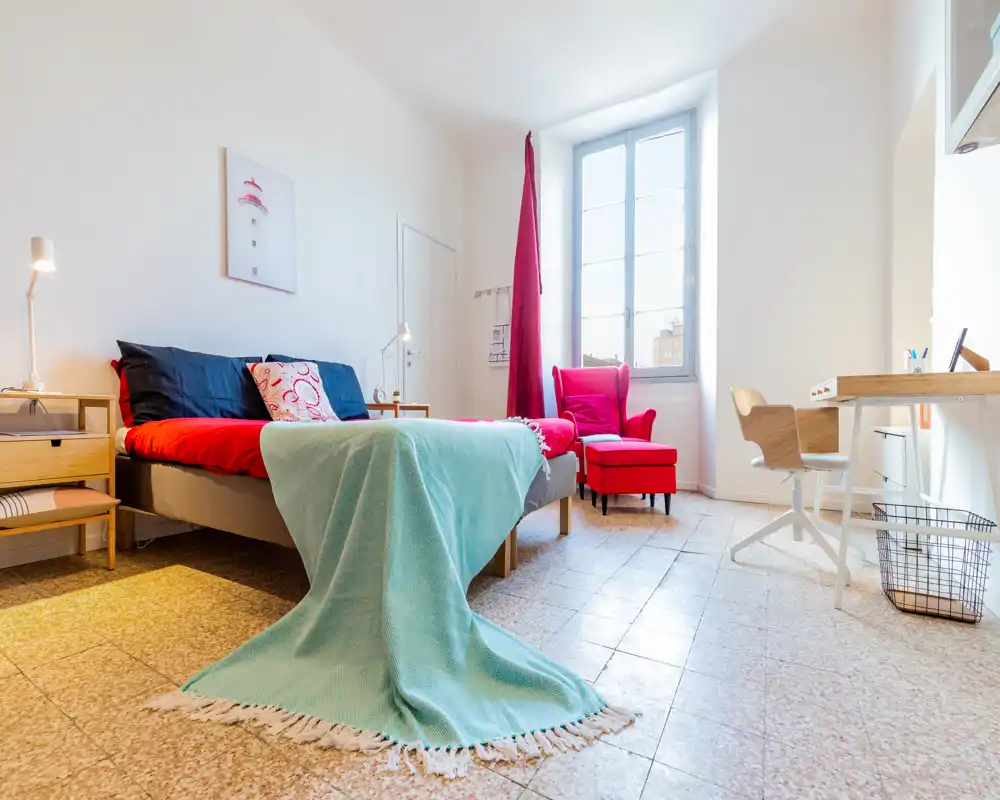 Chambre lumineuse et élégante avec un lit double bien fait, des textiles colorés et un fauteuil confortable près de la fenêtre — idéale pour valoriser le confort et la lumière naturelle.